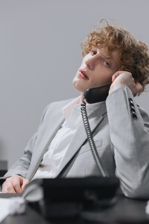 Free Stylish young man in a gray suit talking on a phone at an office desk. Stock Photo