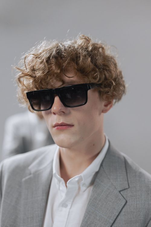 Free Stylish portrait of a young man with sunglasses, curly hair, wearing a gray suit indoors. Stock Photo
