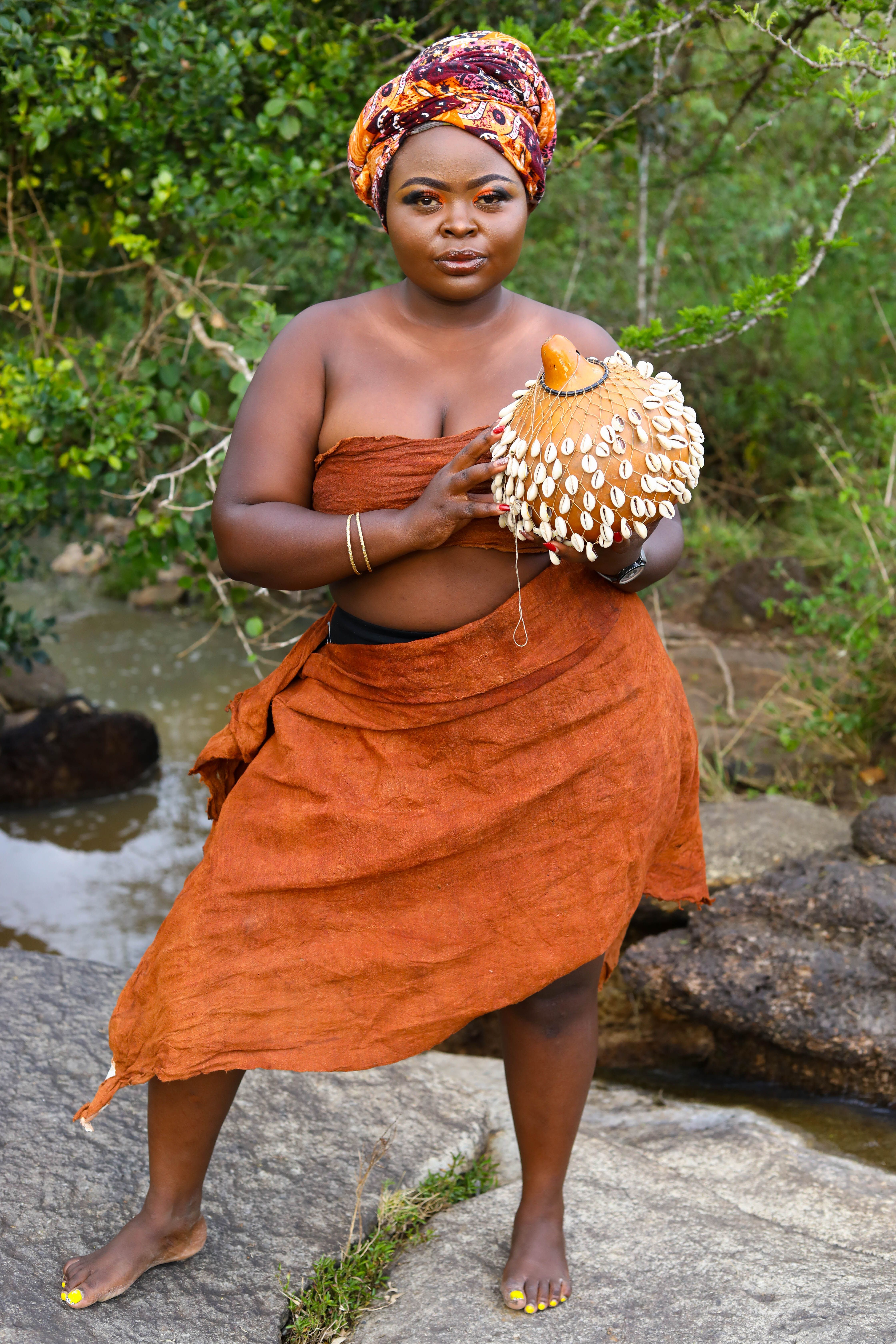 A beautiful portrait of an African woman in traditional dress, barefoot, holding a decorative item, set in a lush outdoor environment.