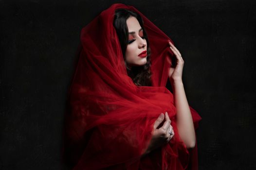 Graceful woman in red veil with closed eyes against black background, highlighting elegance and mystery.