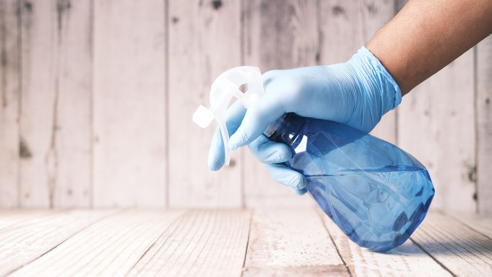 A gloved hand holding a blue spray bottle against a wooden background.