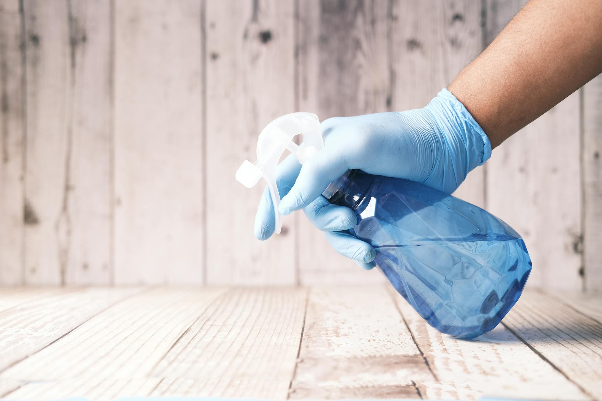 A gloved hand holding a blue spray bottle against a wooden background.