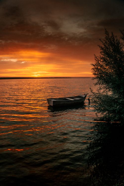 Free A lone boat floats on calm waters with a vibrant orange sunset creating shadows and reflections. Stock Photo