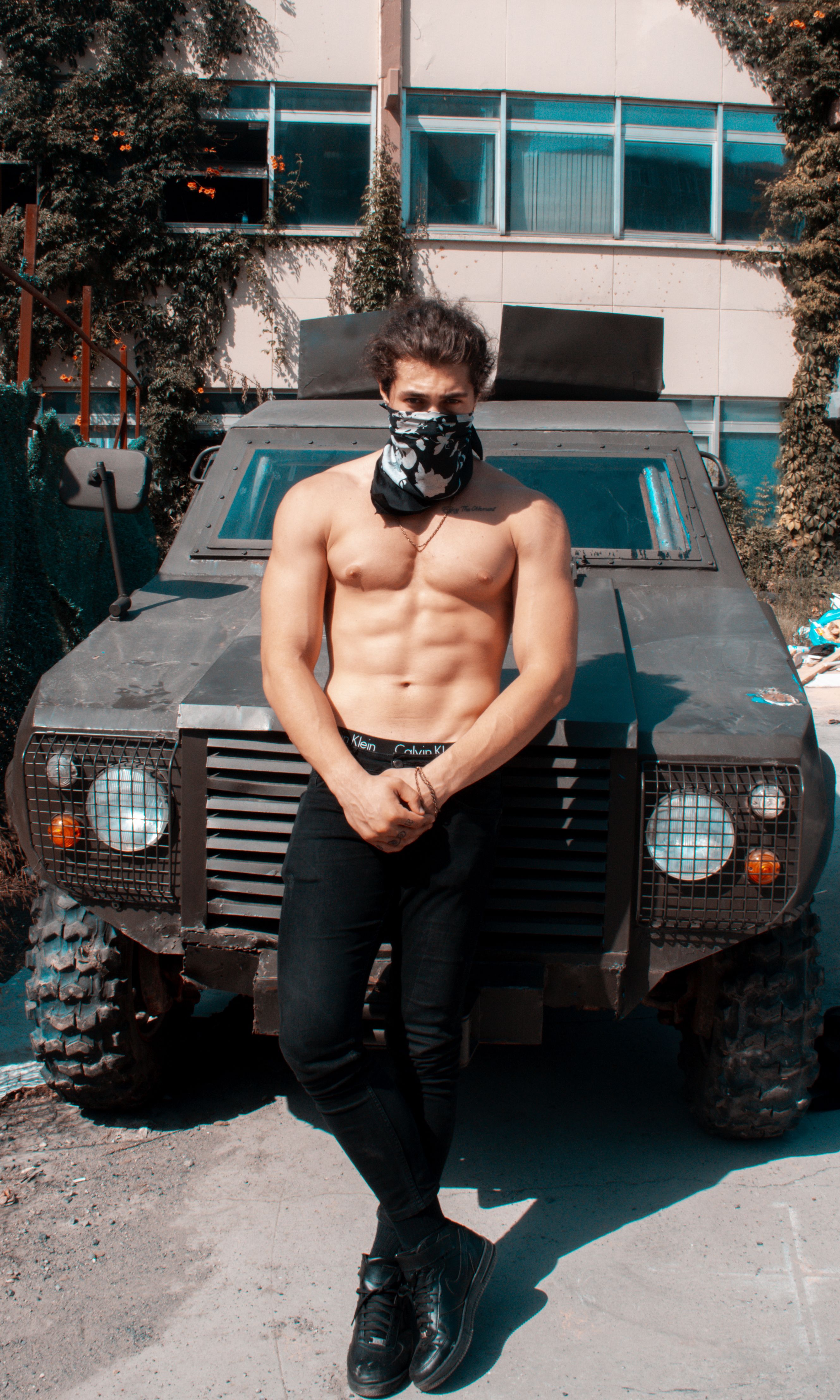 Muscular man wearing a bandana posing confidently against an armored vehicle in an urban setting.