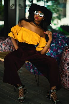Fashionable woman sitting indoors in a chic outfit with hat and sunglasses.