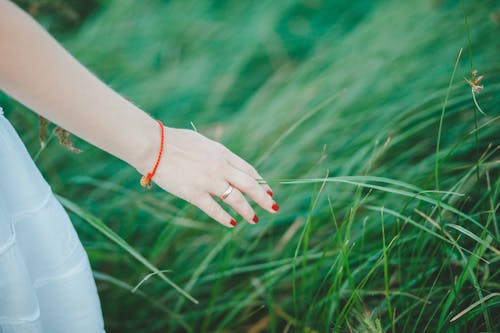 Женщина в белой юбке, оранжевом браслете и красном маникюре у зеленой травы