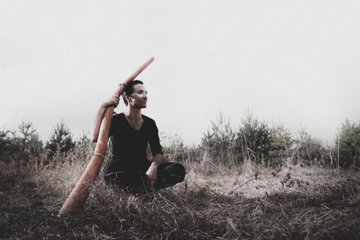 A man seated in a meadow holding a traditional didgeridoo, creating a serene and natural atmosphere.