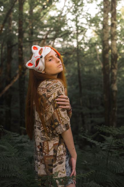Red-haired woman with animal mask posing in lush forest.