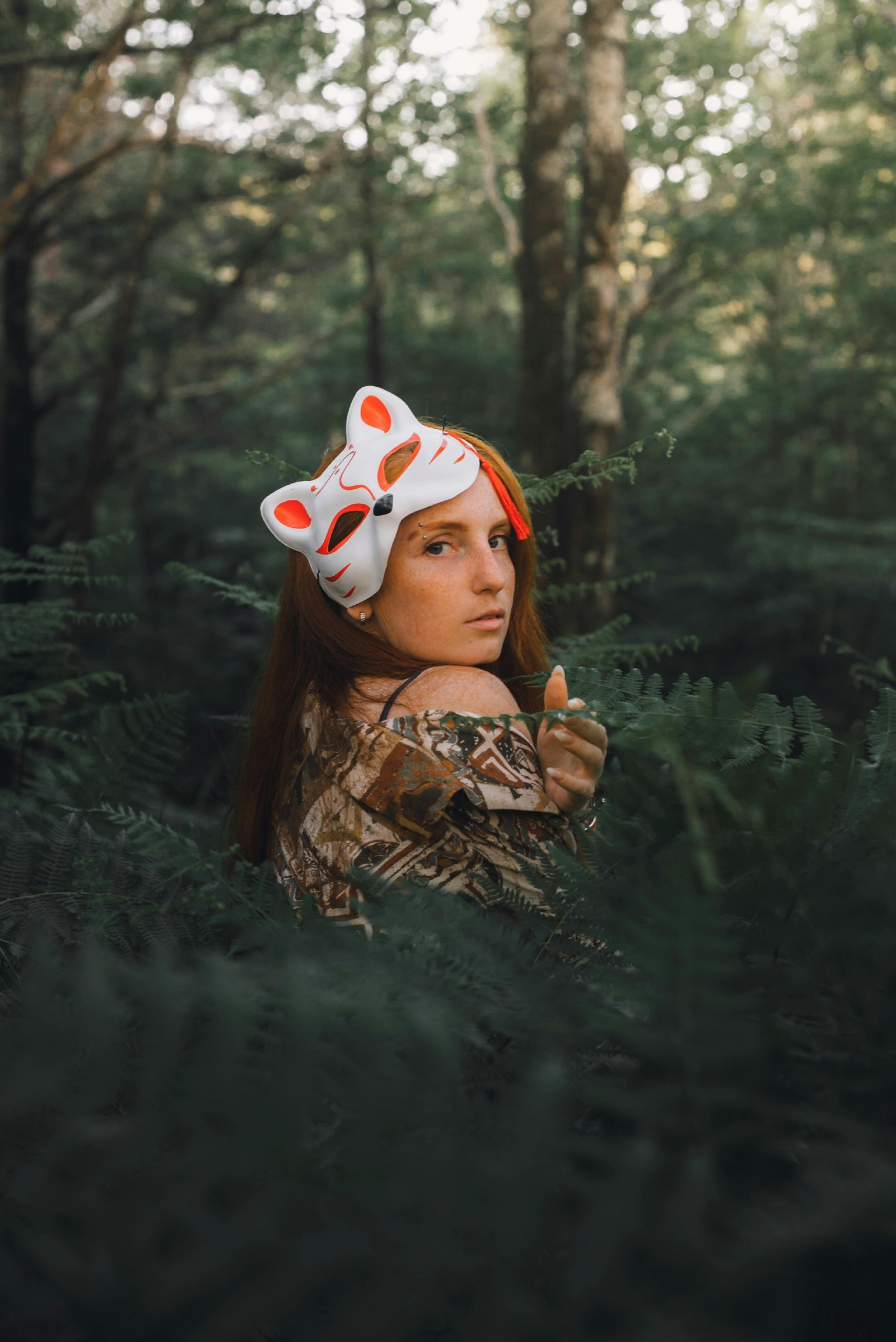 A woman wearing a fox mask poses in a lush green forest, creating a mysterious atmosphere.