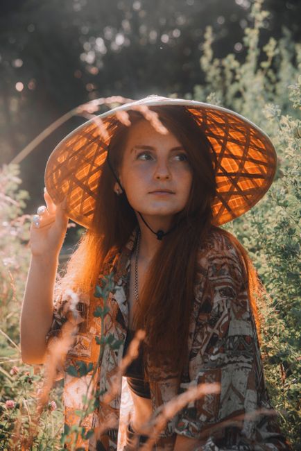 A woman with red hair wearing an Asian conical hat, in a sunny garden.