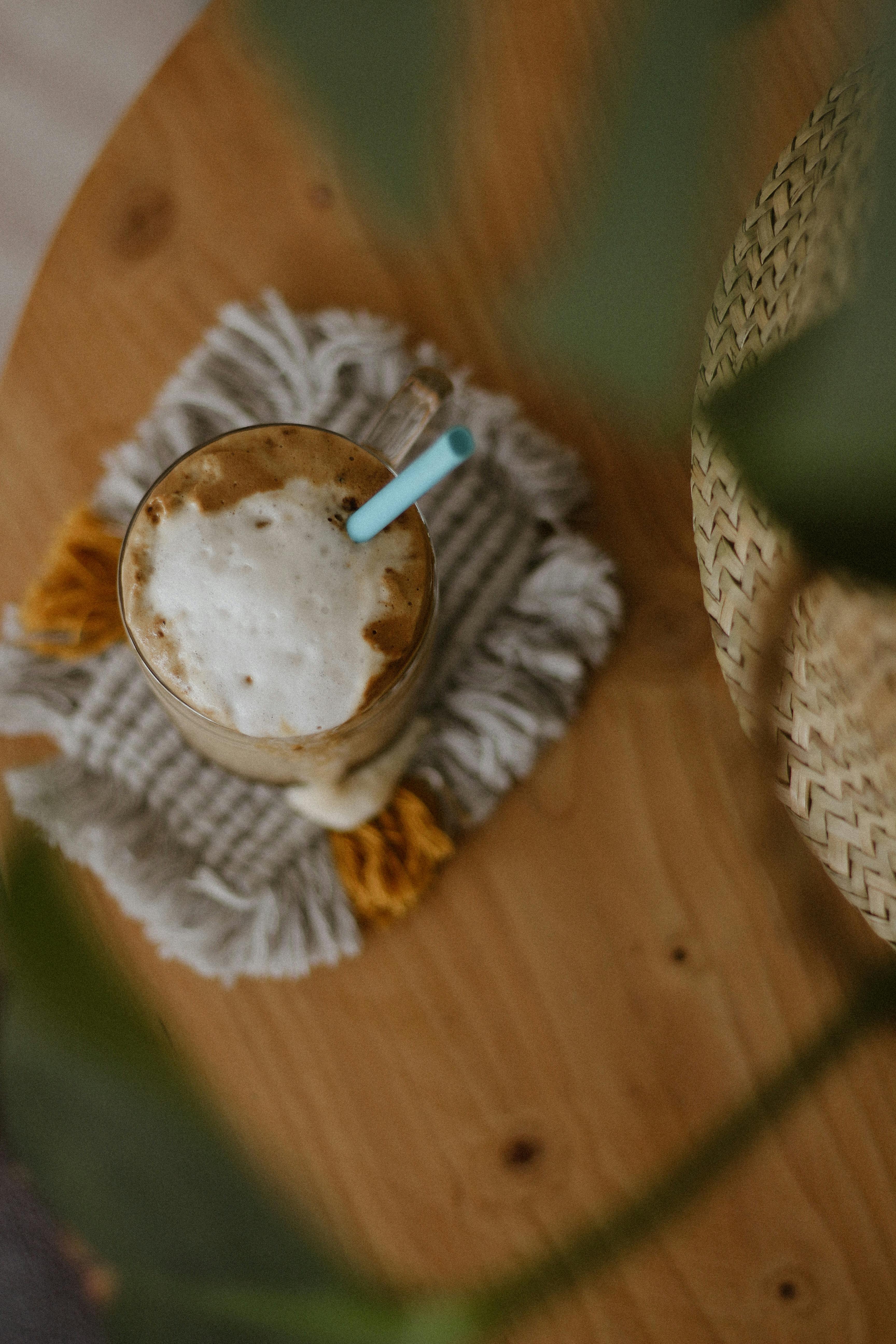 Top view of tasty coffee in glass cup with blue straw placed on knitted coaster near plant on wooden table