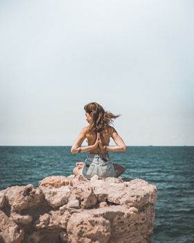 A woman practicing yoga in reverse prayer pose atop rocky shore with ocean background.