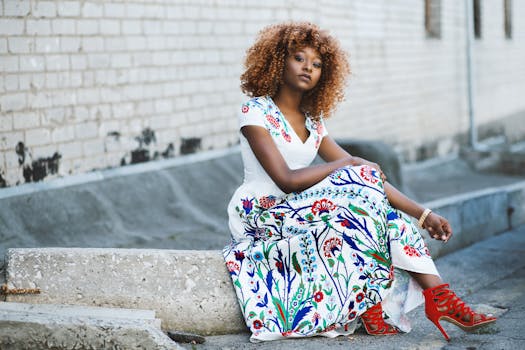 African American woman sitting and posing in a vibrant floral dress against a brick wall.