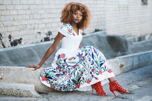 Free Woman in White and Multicolored Floral Flare Dress Sitting on Concrete Stock Photo