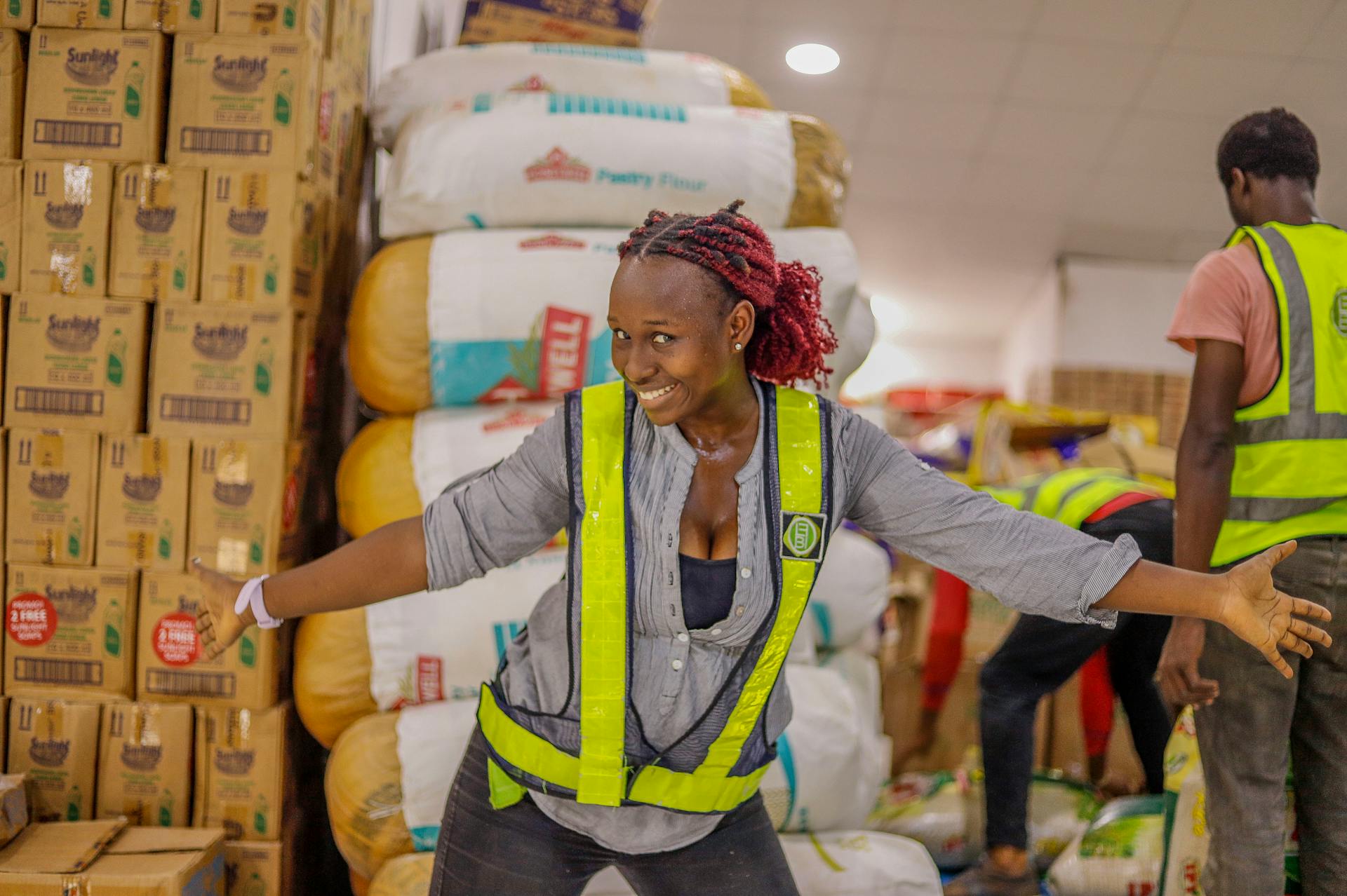 Joyful female worker in a warehouse surrounded by stacked goods, wearing a safety vest.