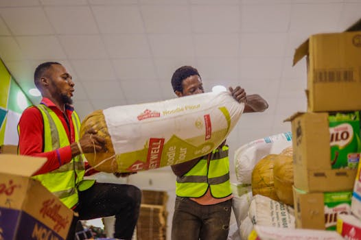 Two male volunteers organizing large sacks and cardboard boxes of relief goods indoors.