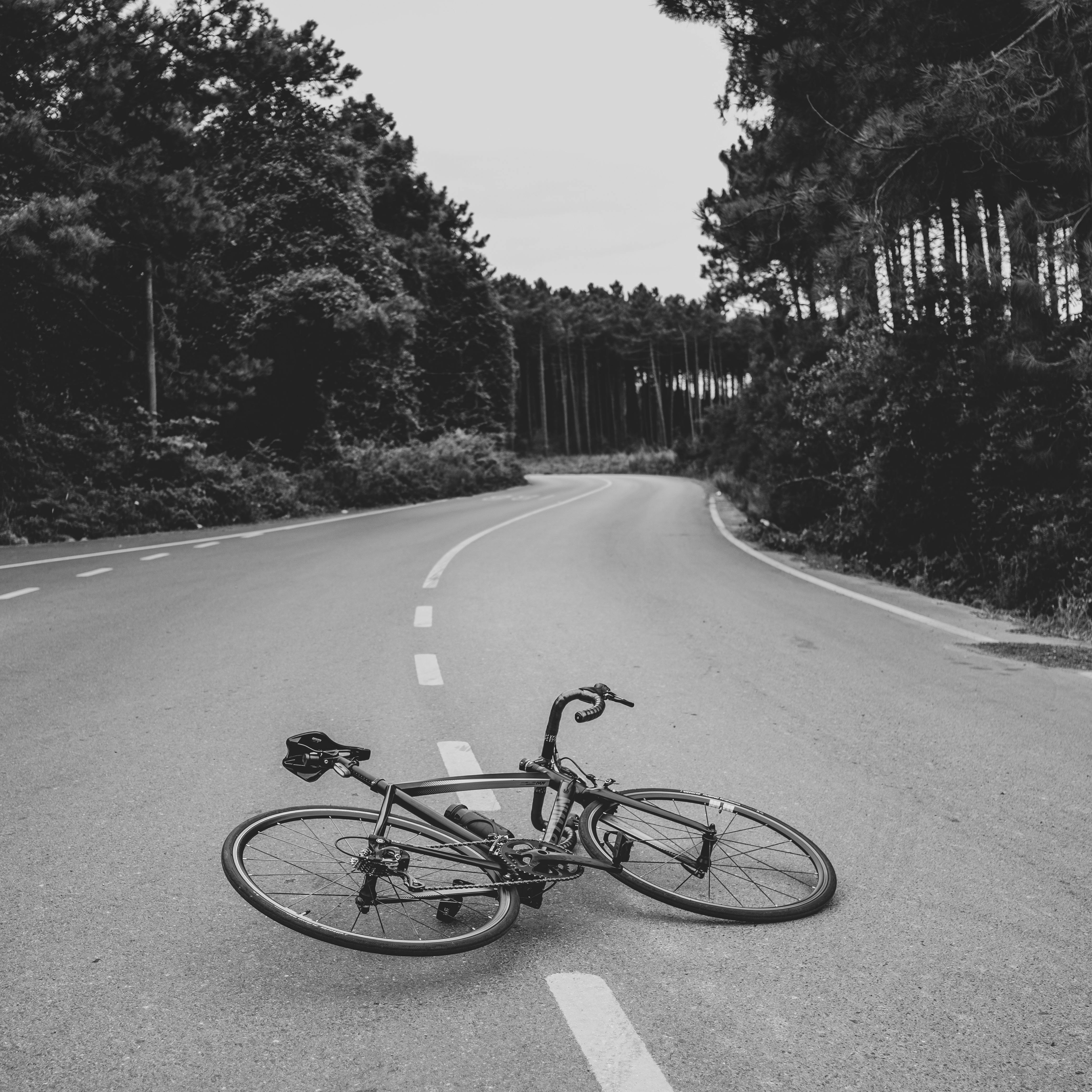 A monochrome image of a bicycle lying on a deserted road surrounded by trees.