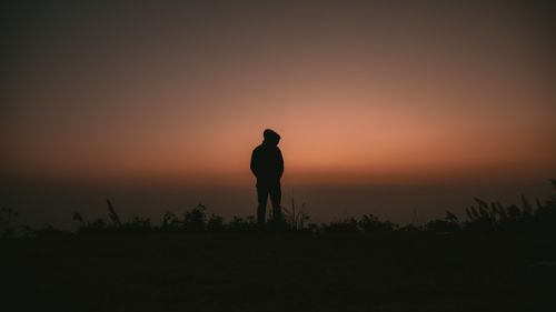 Free Silhouette of a person in a hoodie facing sunset in an open grassy field, evoking solitude and reflection. Stock Photo