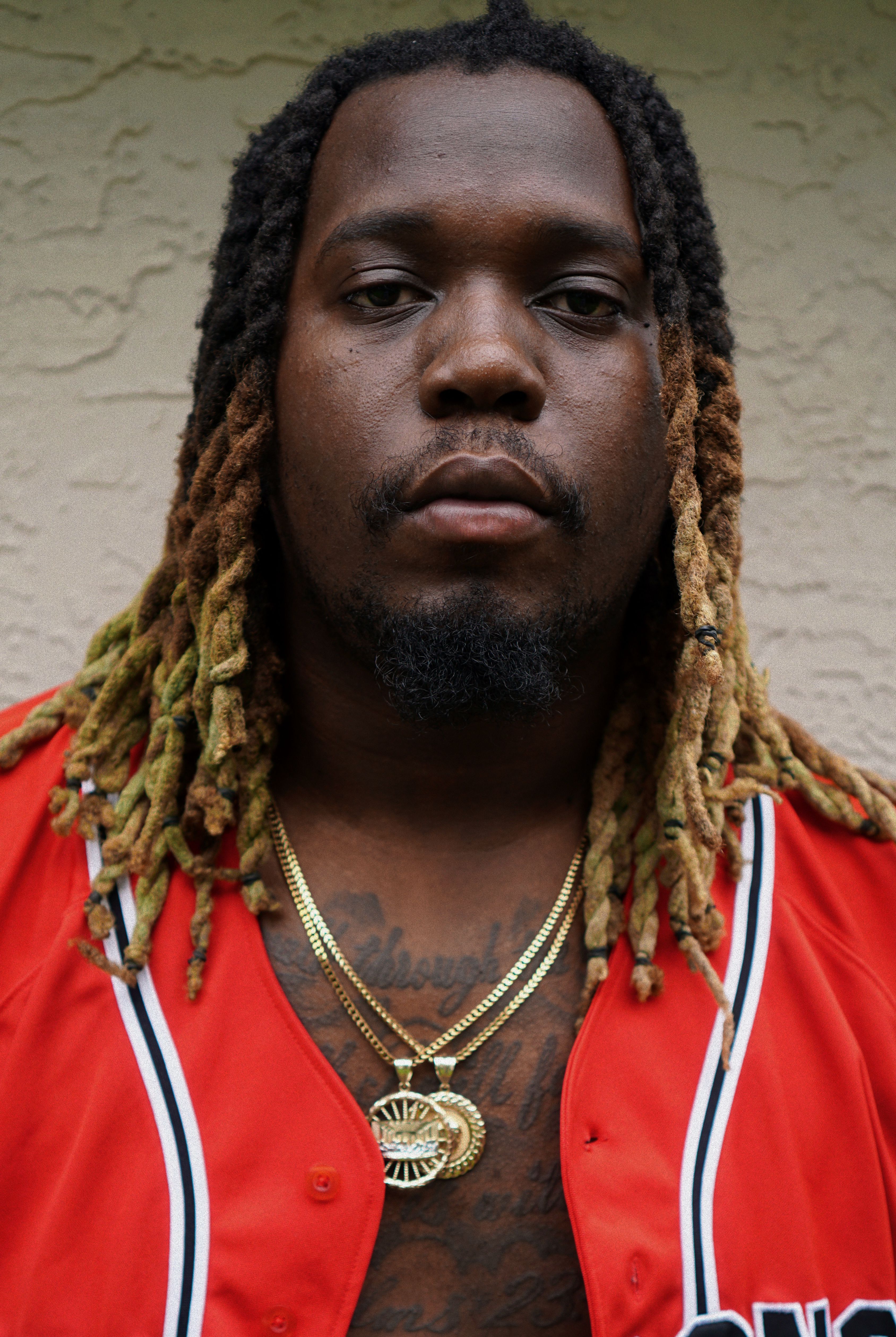 Striking portrait of a man in a red jacket with dreadlocks and gold jewelry, expressing confidence.