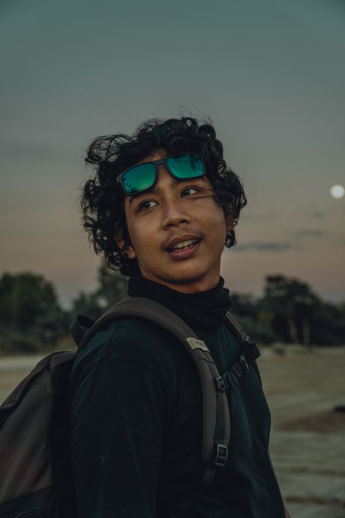 Free Portrait of a young man with curly hair wearing sunglasses on his head, smiling under a warm sunset sky. Stock Photo