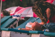 一名年轻女子坐在沙滩椅上，在遮阳伞下用笔记本写字