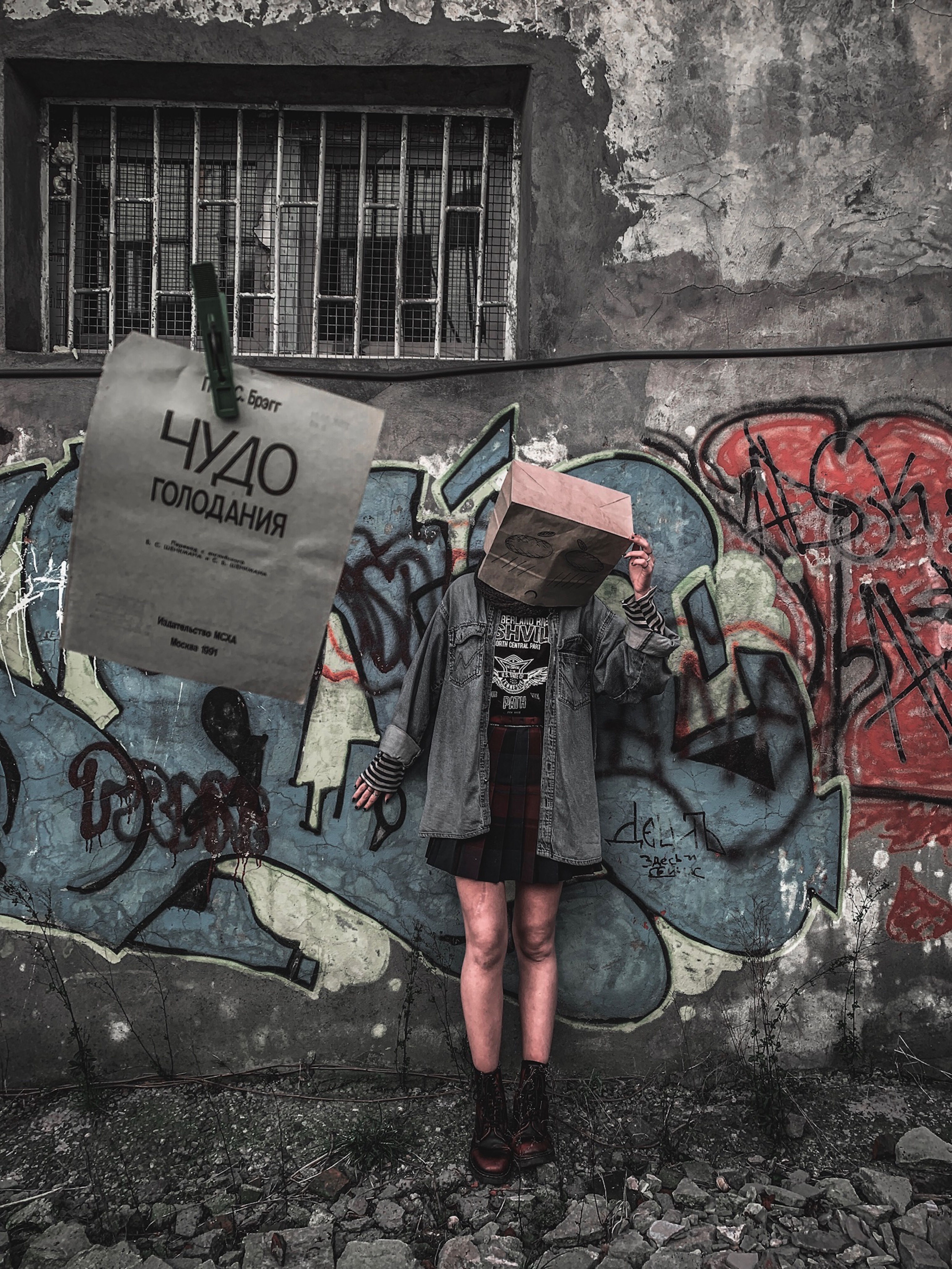 A young woman stands in front of a graffiti wall holding a paper bag over her head, creating an enigmatic urban scene.