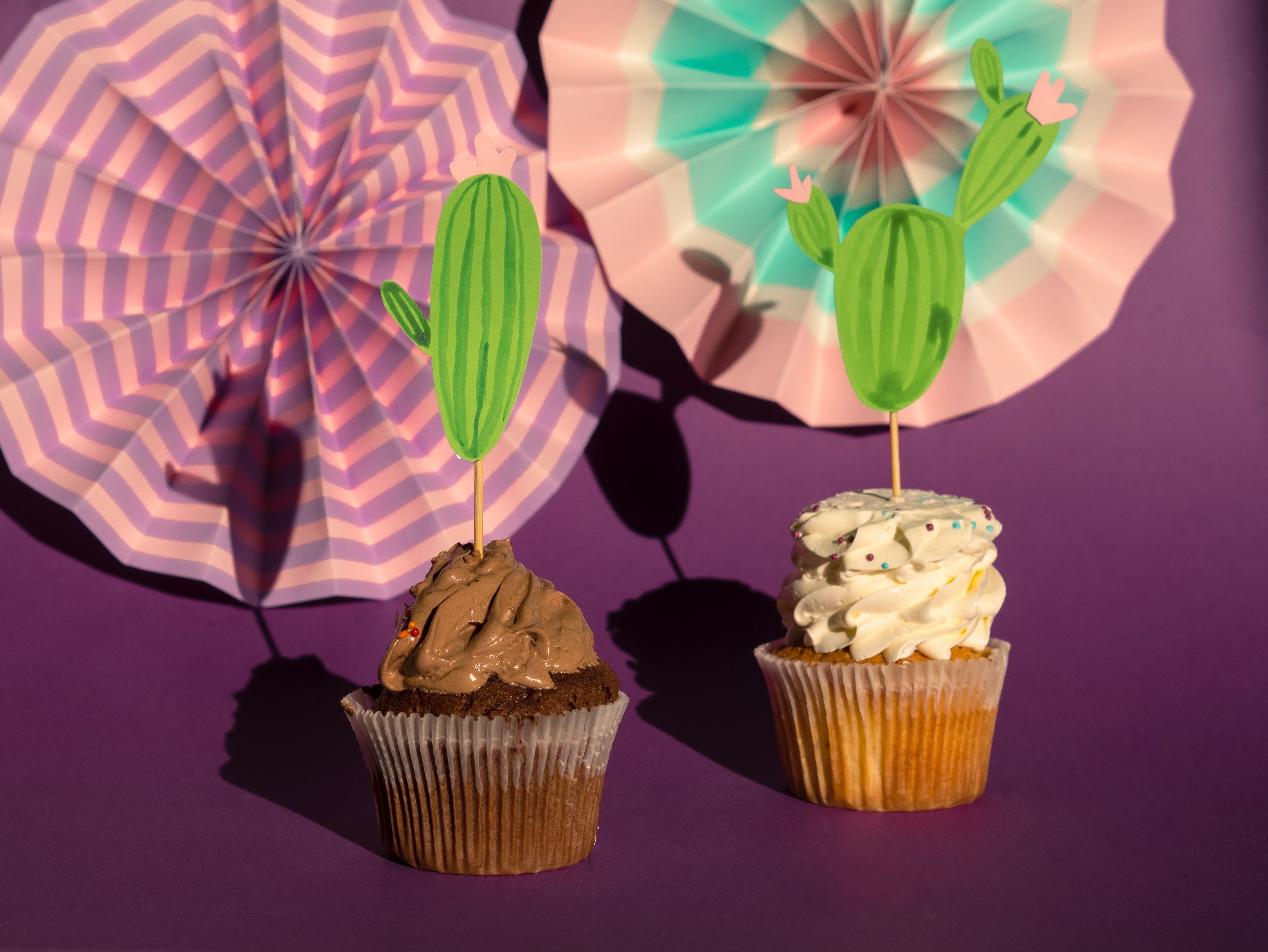 Two cupcakes with cactus toppers against a vibrant purple background with fan decorations.