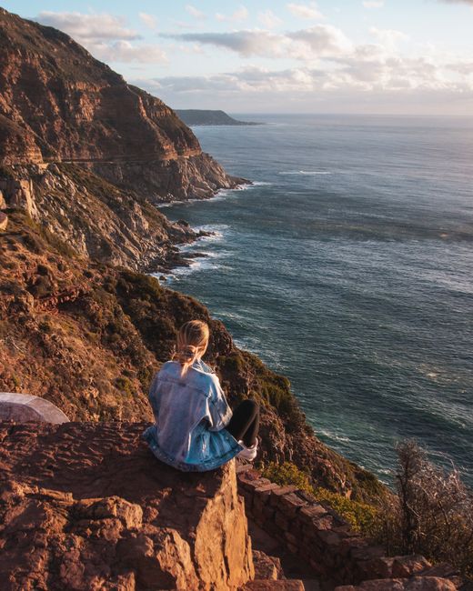 Woman in a denim jacket enjoys a stunning ocean view from Cape Town's rocky cliffs at sunset.