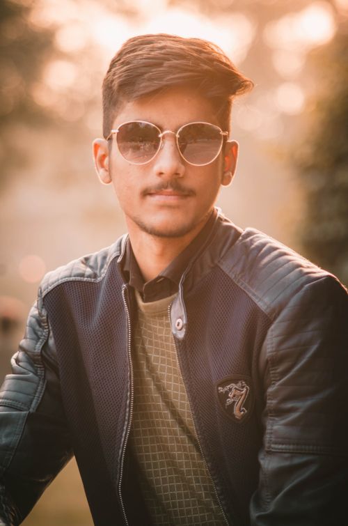 Free Young man posing outdoors with sunglasses, wearing a leather jacket in soft sunlight. Stock Photo
