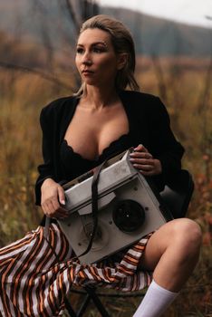 Elegant woman posing outdoors with a vintage reel-to-reel recorder, evoking a retro and fashionable vibe.