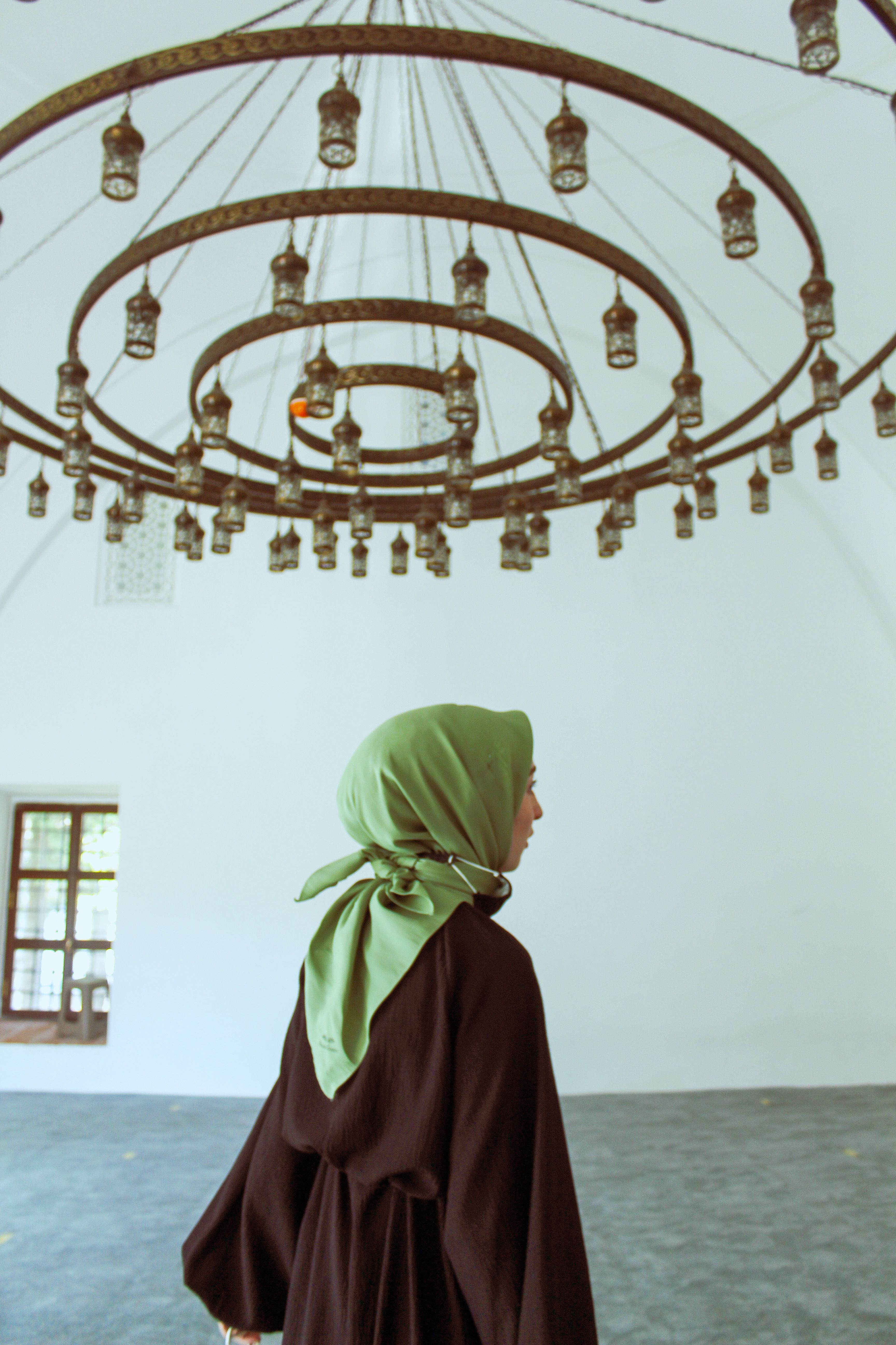 Woman in a green hijab stands beneath an ornate chandelier in a minimal white interior.