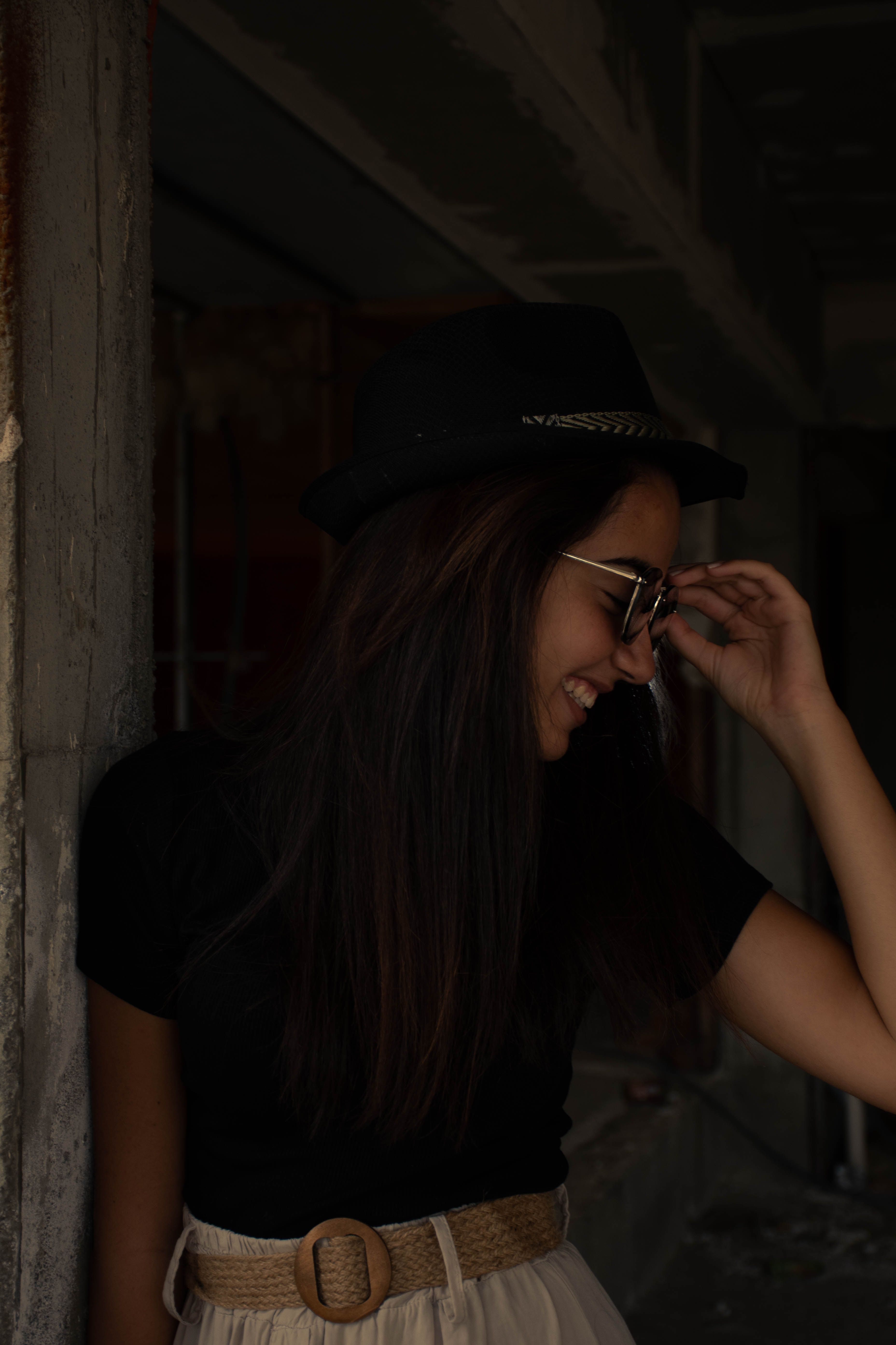 A woman playfully adjusting her hat, smiling gracefully in dim lighting.