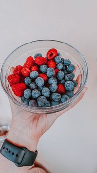A hand holds a glass bowl filled with fresh blueberries and raspberries, offering a vibrant mix of colors.