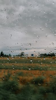 Raindrops on a glass window with a blurred countryside view, creating a moody atmosphere.