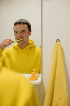 A man in a yellow hoodie brushes his teeth, with a rubber duck and towel in view.