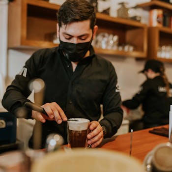 Barista in black uniform frothing milk for coffee in a cozy café environment.