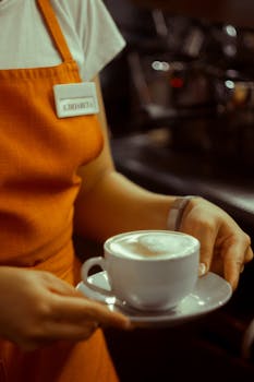 A barista in an apron holds a steaming cup of cappuccino in a cozy café setting.