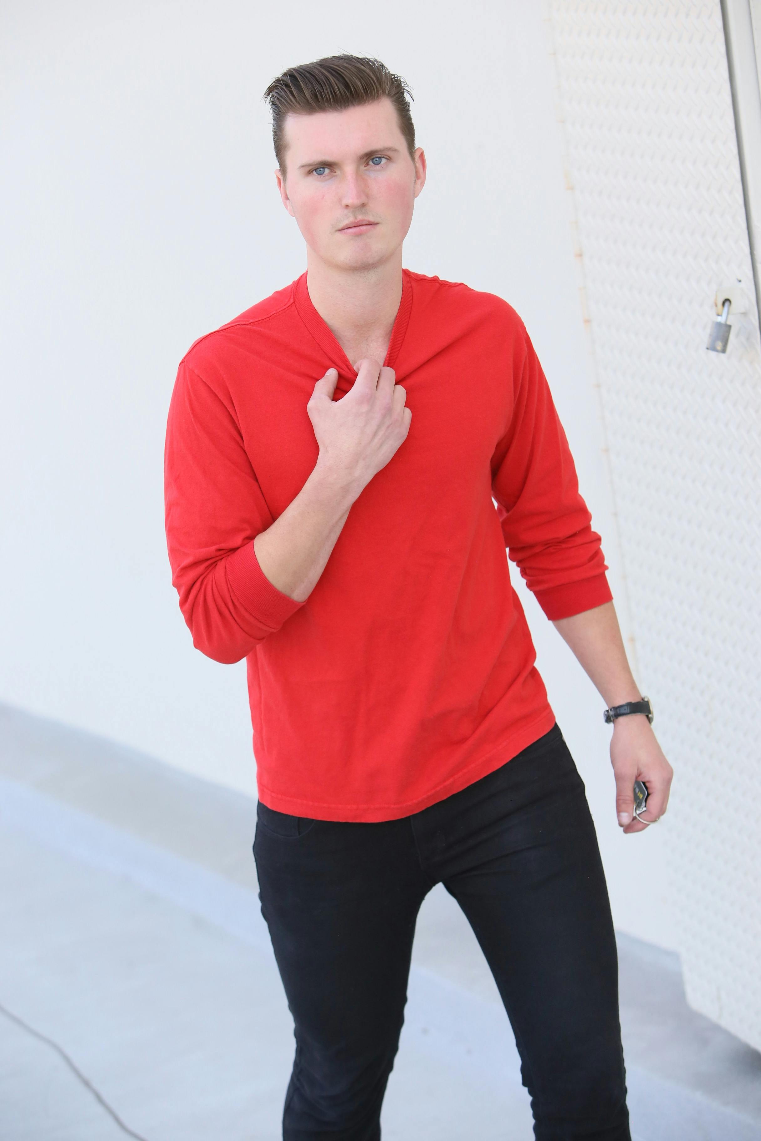 A young man in a red shirt poses confidently against a white wall.