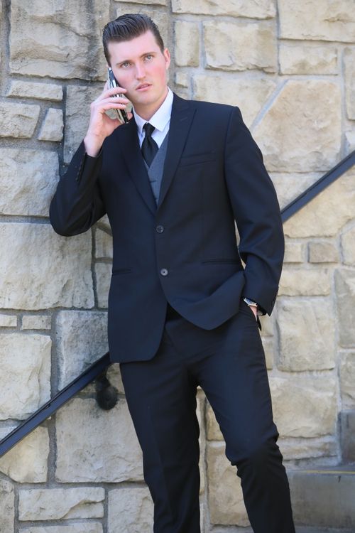 Free Confident businessman in black suit using smartphone outdoors against stone wall. Stock Photo