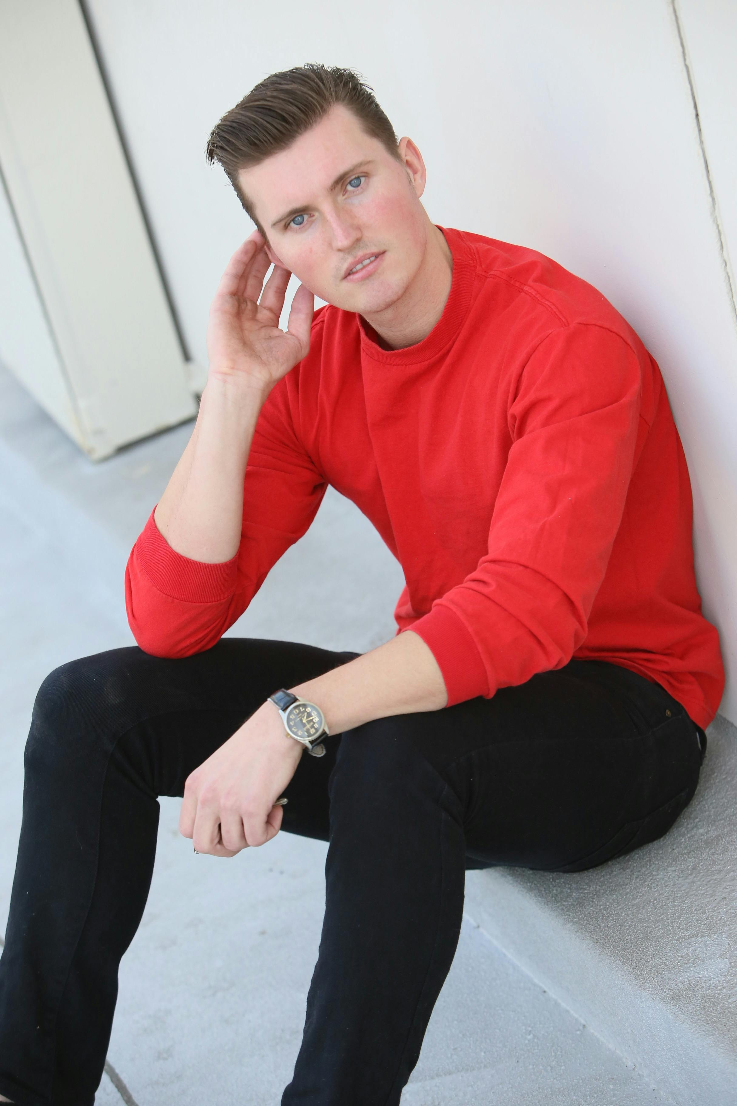 A young man in a red sweater posing casually, seated against a light background.