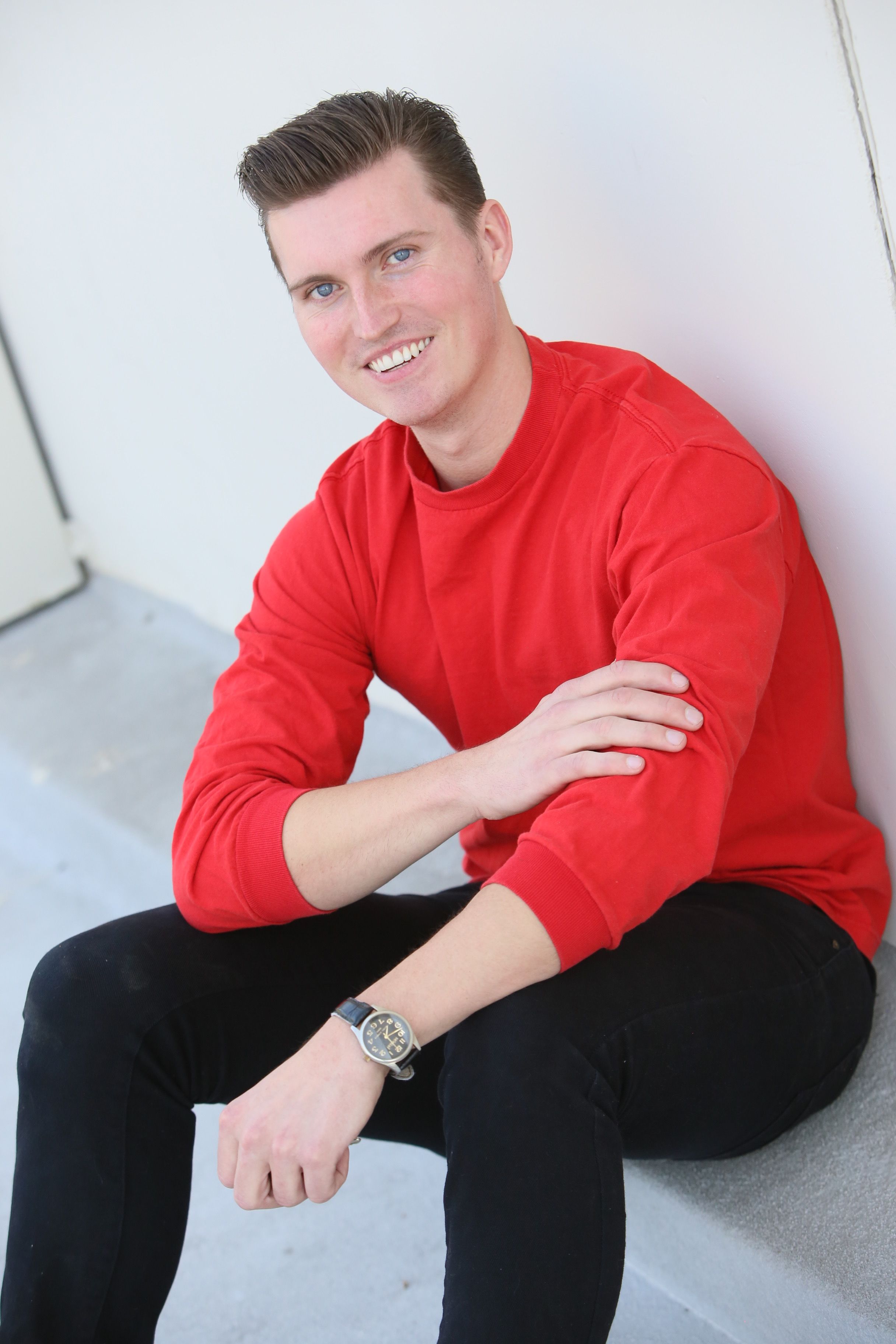 Portrait of a smiling man in a red sweater sitting casually against a wall.