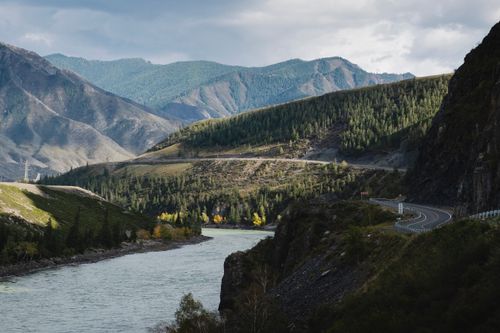 Free Breathtaking view of a winding road alongside a river with majestic mountains in the background. Stock Photo