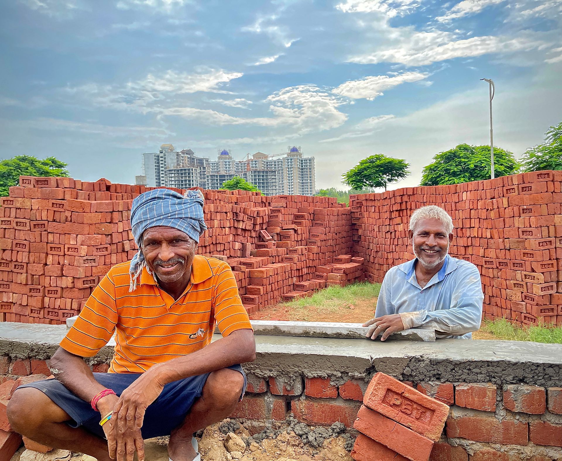 Two smiling workers at an outdoor brick site in Rasulpur, India with urban skyline background.