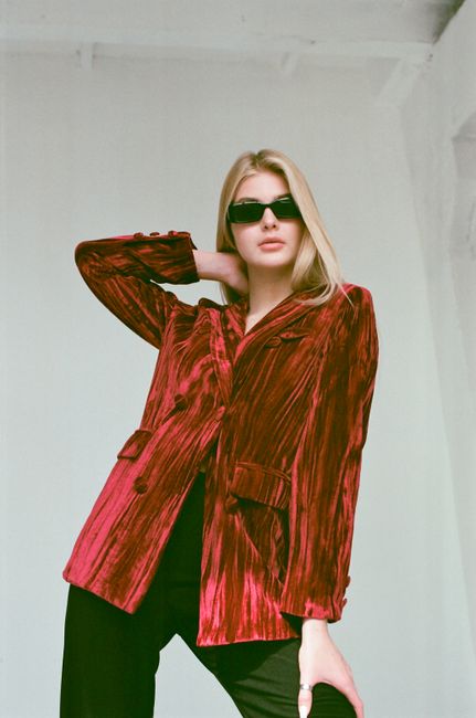 Fashionable woman in red velvet jacket posing indoors with sunglasses.
