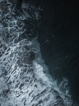 Dramatic aerial shot of ocean waves crashing against the rocky shore, creating a textured seascape.