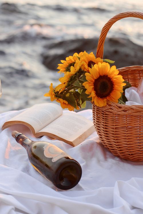 Free A tranquil beach picnic setup with sunflowers, wine, and an open book at sunset. Stock Photo