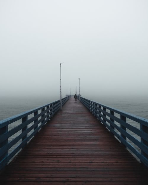 Free A solitary figure walks on a misty wooden pier stretching over the calm ocean. Stock Photo