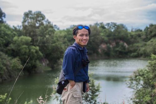 Free A man in blue shirt and sunglasses smiles while posing by a lush green lake during the day. Stock Photo