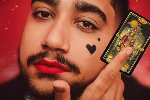 A Man with Heart Shaped Tattoo on His Face Holding a Tarot Card while Looking at the Camera