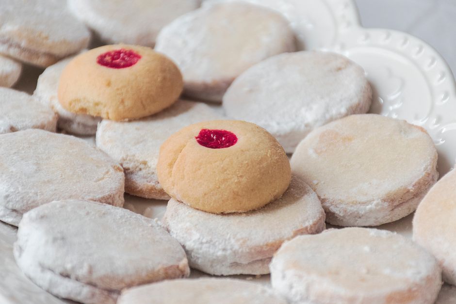 A dozen buttery thumbprint cookies on a wire rack, each filled with a different colored Bonne Maman jam, from deep purple blueberry to bright orange marmalade and pale lemon curd.